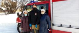 Zdjęcie przedstawia dwie kobiety oraz strażaka OSP Górne stojącego pomiędzy nimi na tle pojazdu pożarniczego, który posłużył jako środek transportu seniorów do punktu szczepień