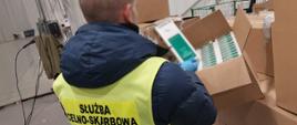 Osoba w kamizelce Służby Celno‑Skarbowej ogląda otwarte pudełko z dużą liczbą paczek papierosów.
