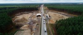 Budowa przejścia górnego dla zwierząt na etapie prac ziemnych. Jedna jezdnia drogi czynna dla ruchu, druga z rozebraną nawierzchnią. Nad nieczynną jezdnią montowana jest konstrukcja z blachy stanowiąca przepust, w którym biegnie jedna jezdnia drogi.
