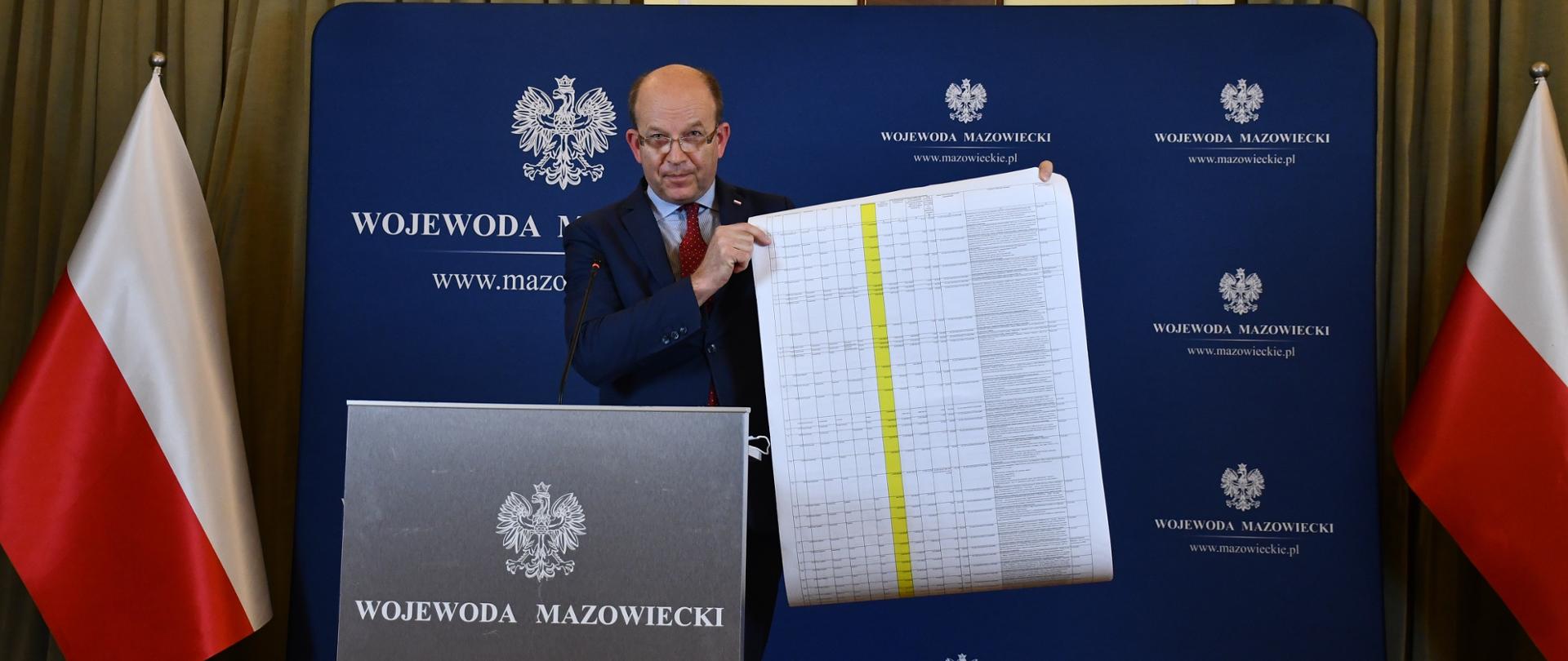 Zdjęcie przedstawia mężczyznę ubranego w garnitur, jasnoniebieską koszulę i czerwony krawat. Mężczyzna ten stoi przed mównicą o stalowym kolorze, na której znajduje się Godło Polski i napis "Wojewoda Mazowiecki". Mężczyzna ten trzyma w rękach długą kartkę papieru, na której znajduje się lista zadań objętych dofinansowaniem w ramach Funduszu. W tle biało-czerwone flagi, ścianka wizerunkowa Wojewody Mazowieckiego oraz znajdujące się na ścianie Godło Polski.