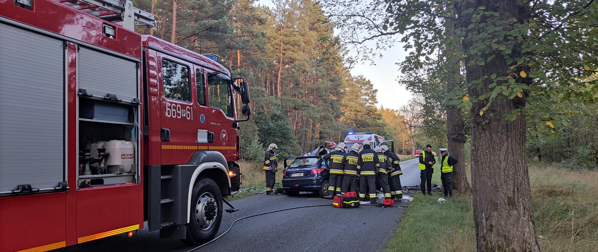 Na zdjęciu widoczny jest pojazd strażacki oraz samochód osobowy przy którym strażacy udzielają pomocy osobom poszkodowanym