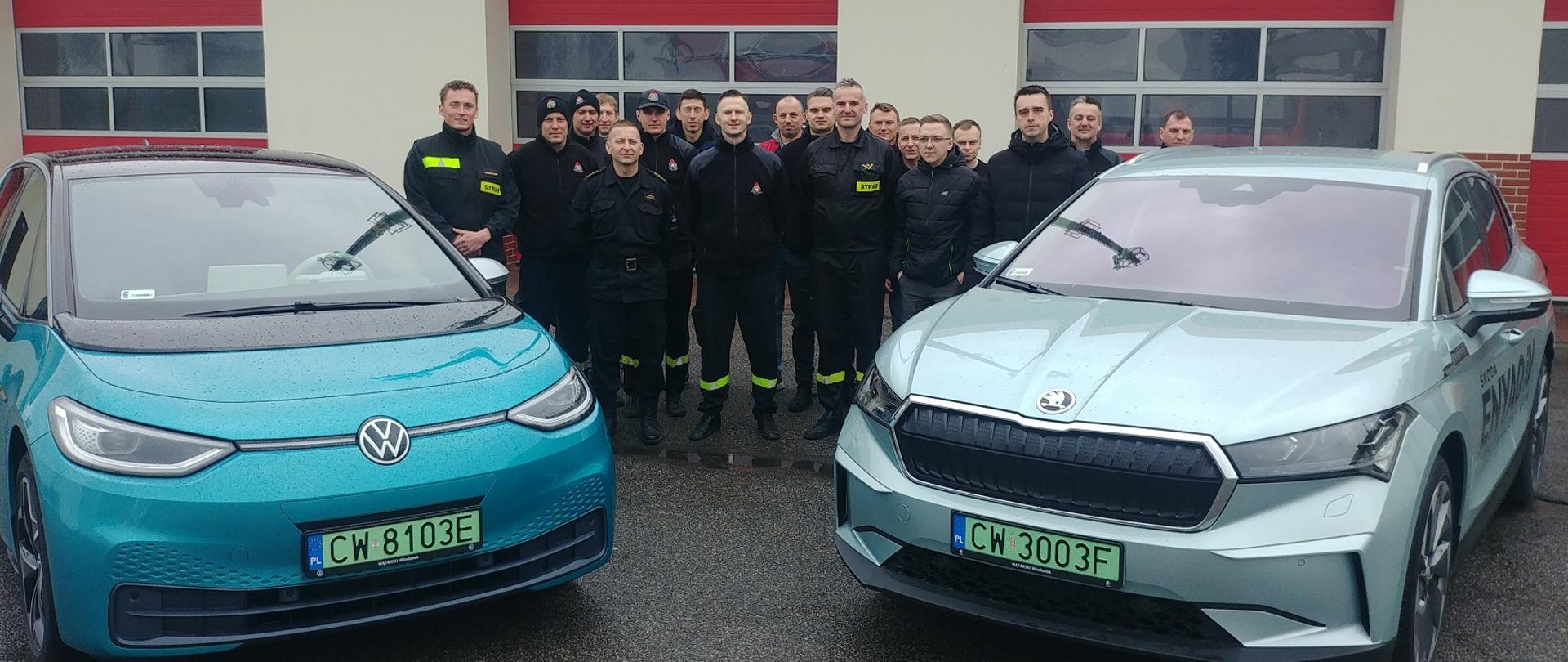 Zdjęcie przedstawia na pierwszym planie dwa samochody elektryczne marki skoda oraz wolkswagen, na drugim planie strażacy Państwowej Straży Pożarnej 