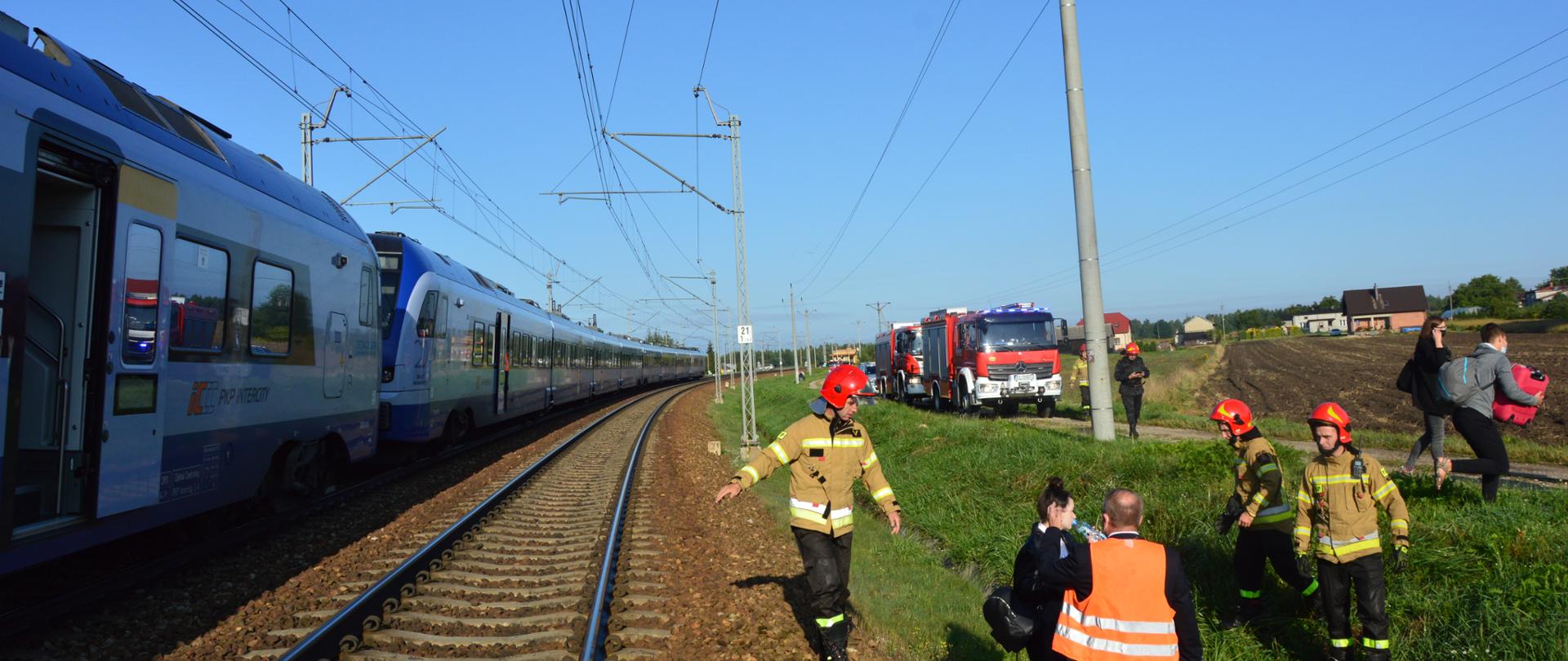 zdjęcie przedstawia strażaków JRG Koluszki na torowisku kolejowym , w oddali widoczne samochody pożarnicze, z lewej pociąg PKP Intercity. 