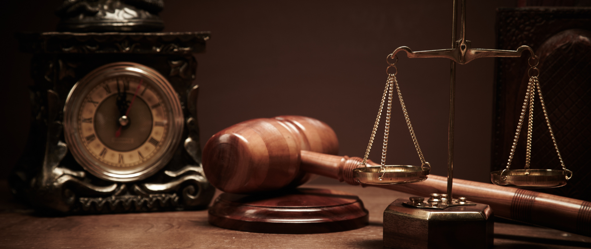 Na zdjęciu: Blat biurka. Na nim drewniany młotek sędziowski na podstawce, ozdobna waga i drewniany zegar. 