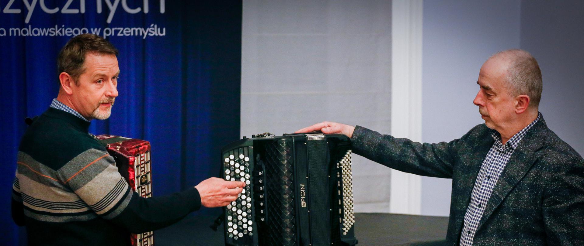 Na zdjęciu widocznych jest dwóch mężczyzn prezentujących akordeony. Jeden z nich trzyma czerwony instrument i wskazuje na klawiaturę guzikową, natomiast drugi stoi obok i opiera rękę na czarnym akordeonie ustawionym na stole. Spotkanie odbywa się w sali, a w tle widać fragment sceny z kurtyną.