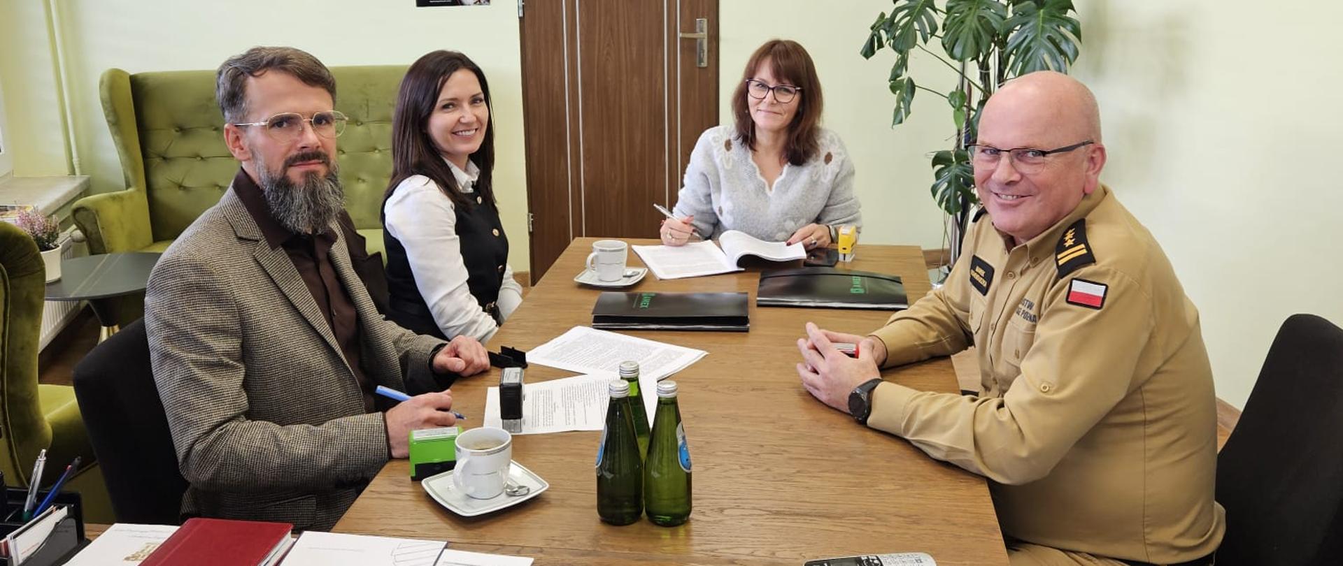 Cztery osoby siedzą przy stole konferencyjnym w biurze. Po lewej stronie mężczyzna w marynarce, obok niego dwie kobiety, a po prawej stronie mężczyzna w piaskowym mundurze strażackim. Wszyscy uśmiechają się w stronę kamery. Na stole znajdują się dokumenty, filiżanki i woda mineralna. W tle widać roślinę doniczkową, kalendarz, krzyż i ikonę.