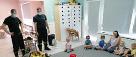 Zdjęcie przedstawia dzieci z Akademii Wiedzy i Zabawy wraz z opiekunami oraz strażakami, którzy w sali zajęć opowiadają o pracy strażaka oraz prezentują hełm strażacki i ubranie specjalne nomeks.