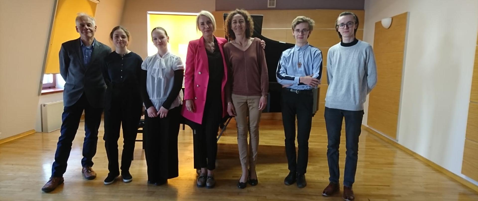 Uczestnicy warsztatów pianistycznych na sali kameralnej stoją w zwartym szyku. Za nimi fortepiany oraz dwa okna przysłonięte roletą.