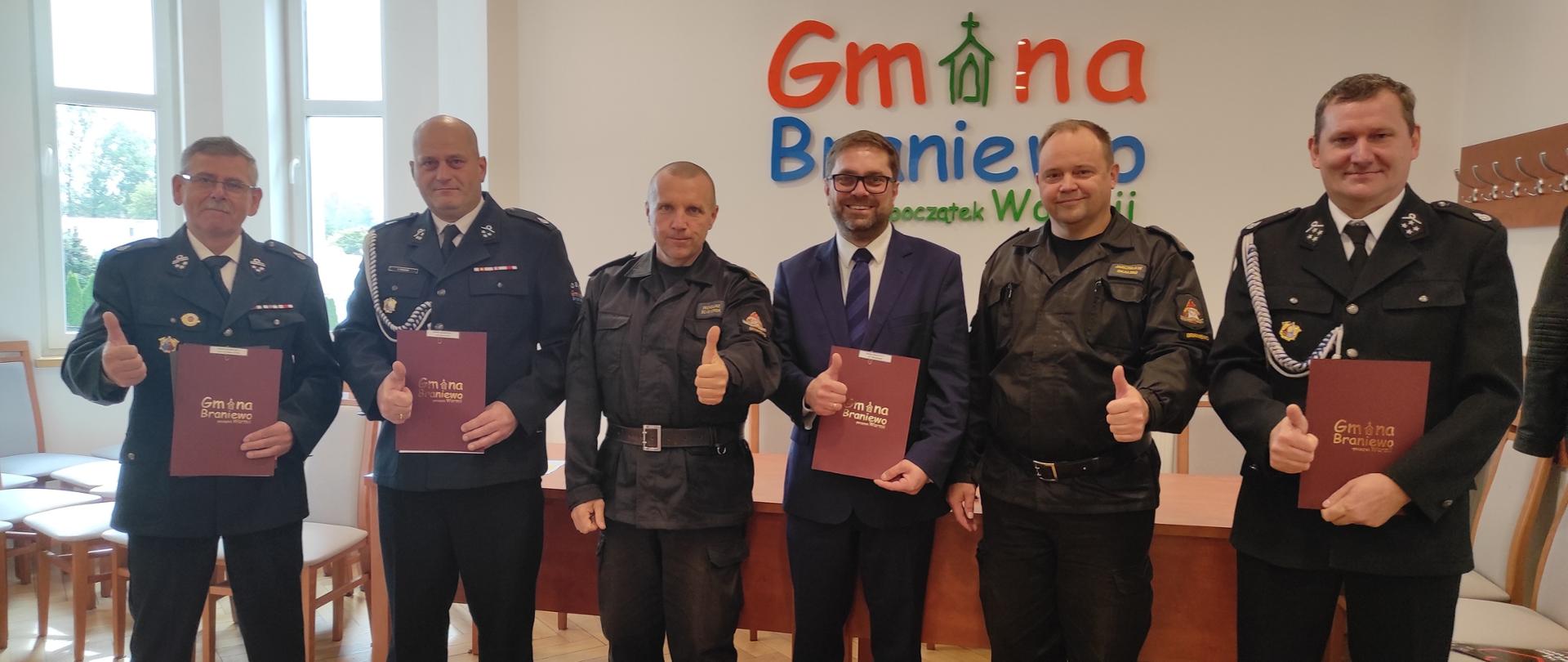 Na tle ścianki z napisem Gmina Braniewo stoją ustawieni obok siebie sześć osób, strażacy w mundurach oraz wójt w garniturze. 