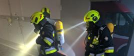 Zdjęcie przedstawia trzech strażaków-ratowników, którzy pełnią funkcję roty ratowniczej RIT. Są oni ubrani w pełne ubranie specjalne wraz z wymaganym wyposażeniem czyli: aparatem ochrony układu oddechowego, latarką zamontowaną przy hełmie, kamerą termowizyjną oraz miernikiem, służącym do monitorowania atmosfery w której się znajdują. Ratownicy ewakuują czwartego strażaka - poszkodowanego, który znajduje się w pozycji leżącej. Jest on również ubrany w ubranie specjalne. Sytuacja ma miejsce w garażu, w którym występuje zadymienie ograniczające widoczność ratownikom. Cała sytuacja ma miejsce obok drzwi, w których widać czerwony samochód. Po lewej stronie widać leżące na ziemi przewody elektryczne, które mogą utrudnić strażakom ewakuację