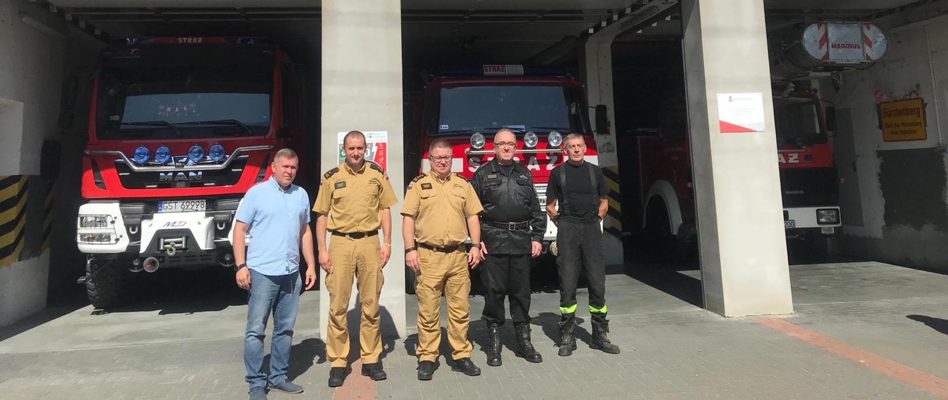 Zdjęcie przedstawia wizytujące osoby na tle jednostki OSP. W tle widać garaże a w nich samochody pożarnicze