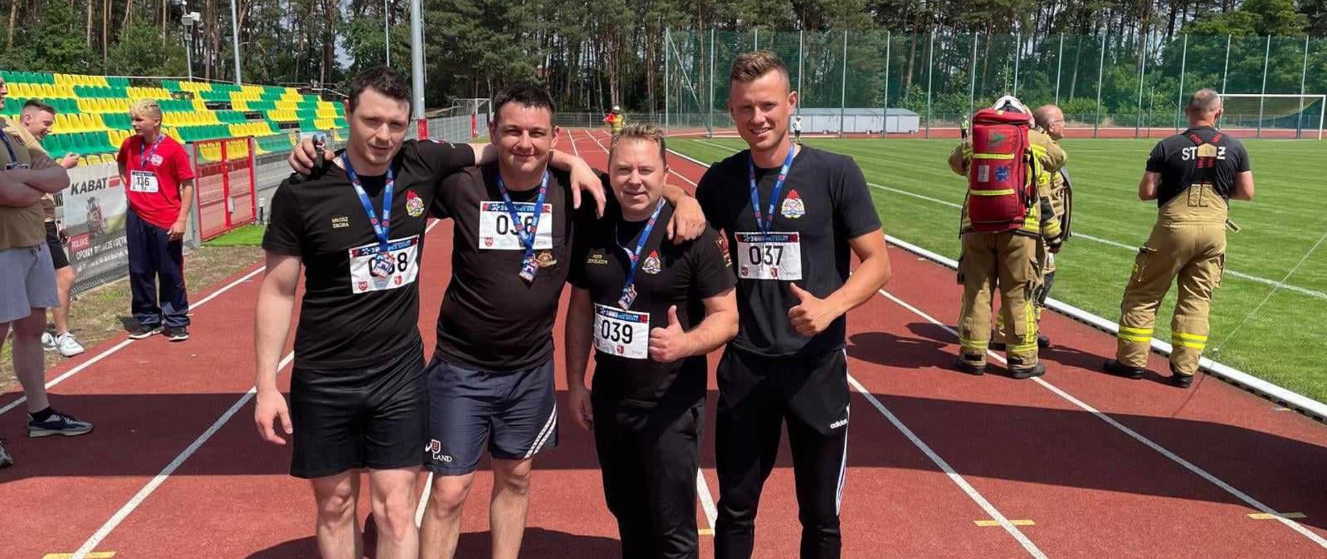 Zdjęcie przedstawia strażaków w trakcie zawodów sportowych.
W tle stadion sportowy.
