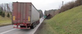 Droga o przebiegu jednojezdniowym wskazująca na wjazd do miejscowości Olsztyn. Po drodze przejeżdża czerwona ciężarówka. Po prawej stronie za znakiem mała drewniana chatka.