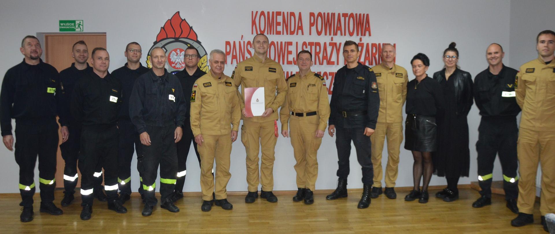 Wręczenie listu gratulacyjnego. Zdjęcie przedstawia świetlicę komendy. Na tle ściany, na której znajduje się logo PSP i czerwony napis KOMENDA POWIATOWA PAŃSTWOWEJ STRAŻY POŻARNEJ W RAWICZU do wspólnego zdjęcia pozują strażak - złoty medalista wraz z kierownictwem komendy oraz strażakami i pracownicą cywilną. Strażacy występuja w mundurach koloru piaskowego oraz czarnego. Strażak-sportowiec trzyma w rękach teczkę z listem gratulacyjnym.