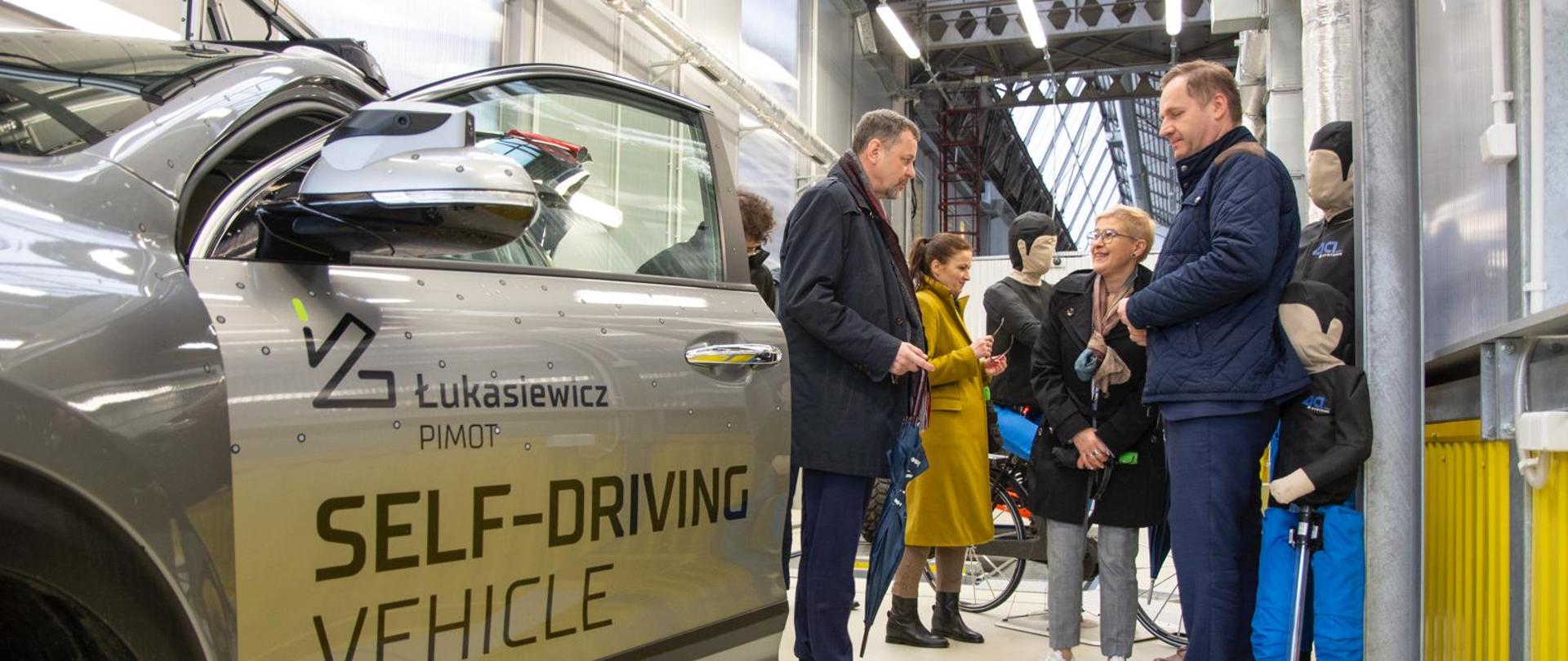 W korytarzu przy szarym samochodzie z napisem na drzwiach Self-driving vehicle stoi grupa osób.