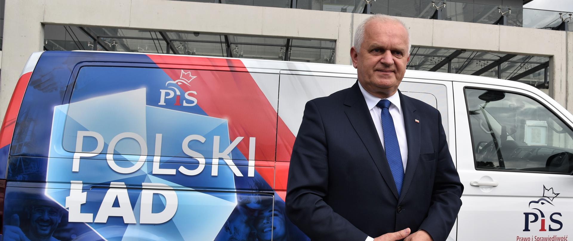 Wojewoda stoi na tle busa z granatowo-czerwoną grafiką i napisem "Polski Ład". W tle niebieski budynek Lubuskiego Urzędu Wojewódzkiego 