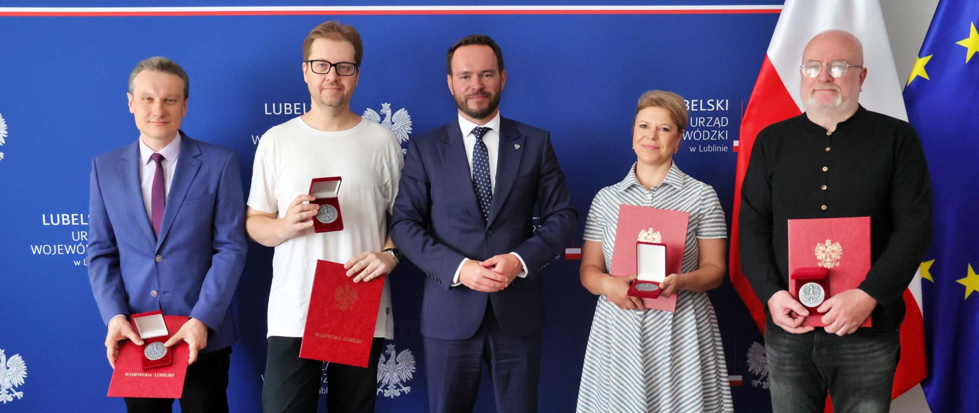 Pięć osób, w tym Wojewoda Lubelski Krzysztof Komorski (czwarty od lewej, w granatowym garniturze), pozuje do zdjęcia na tle flag Polski i Unii Europejskiej z okazji 45-lecia czasopisma „Akcent”. Na zdjęciu są również wyróżnieni laureaci, którzy trzymają czerwone teczki z dokumentami i metalowe medale w etui. Scena ma miejsce w Lubelskim Urzędzie Wojewódzkim.
