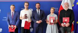 Pięć osób, w tym Wojewoda Lubelski Krzysztof Komorski (czwarty od lewej, w granatowym garniturze), pozuje do zdjęcia na tle flag Polski i Unii Europejskiej z okazji 45-lecia czasopisma „Akcent”. Na zdjęciu są również wyróżnieni laureaci, którzy trzymają czerwone teczki z dokumentami i metalowe medale w etui. Scena ma miejsce w Lubelskim Urzędzie Wojewódzkim.