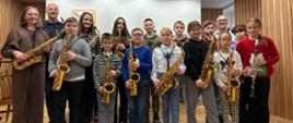 Pionowe zdjęcie grupowe przedstawia grupę młodych muzyków, trzymających instrumenty dęte, w otoczeniu dwóch dorosłych. Większość dzieci trzyma saksofony, a jedno klarnet, stojąc w jasnym, nowoczesnym pomieszczeniu z drewnianymi panelami na ścianach.