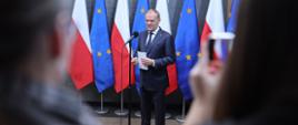 Premier Donald Tusk podczas oświadczenia przed wylotem na szczyt Rady Europejskiej 20.03.2025. W tle flagi Polski i Unii Europejskiej.