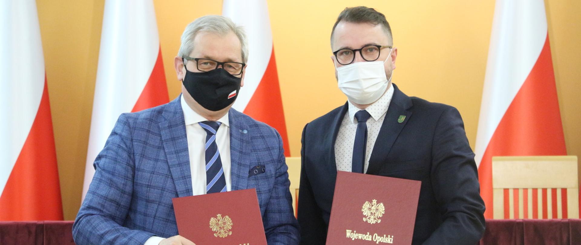 Na zdjęciu wojewoda opolski wraz z wójtem gminy Łubniany, stoją na tle biało-czerwonych flag. W dłoniach trzymają po jednej czerwonej teczce. 
