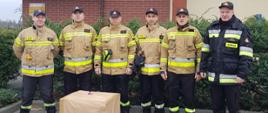 Zdjęcie przedstawia 6 strażaków PSP stojących na tle budynku Komendy Powiatowej Państwowej Straży Pożarnej w Rypinie. Strażacy stoją za dużą paczką. 5 strażaków od lewej ubranych jest w czapki, piaskową górę nomexu oraz spodnie koszarowe. Pierwszy strażak od prawej ubrany jest w czapkę oraz ubranie dowódczo - sztabowe. 