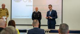 Wojewoda i dwóch strażaków stoją na tle ekranu z prezentacją 