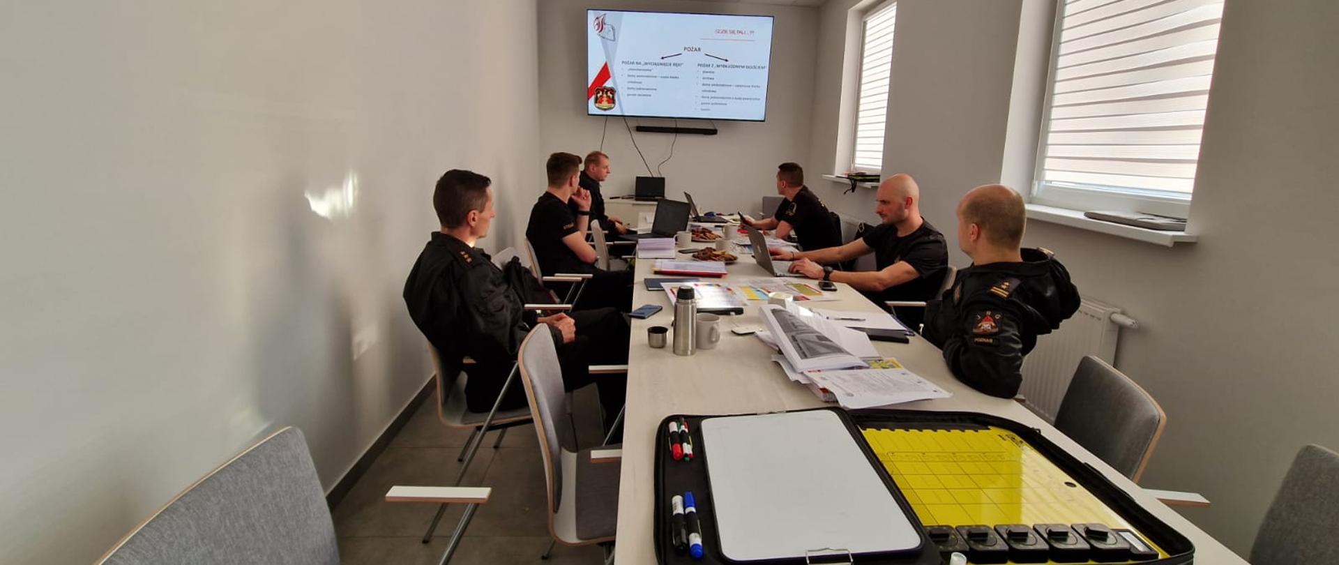 Strażacy siedzą przy stole w pomieszczeniu, w tle prezentacja.