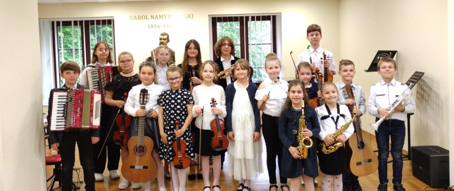 Na pierwszym planie zdjęcia znajduje się grupa 17 uczniów w strojach galowych, z różnymi instrumentami w dłoniach. W tle widoczne popiersie Karola Namysłowskiego, dwa okna, ściany białe, na suficie kilka lapm.