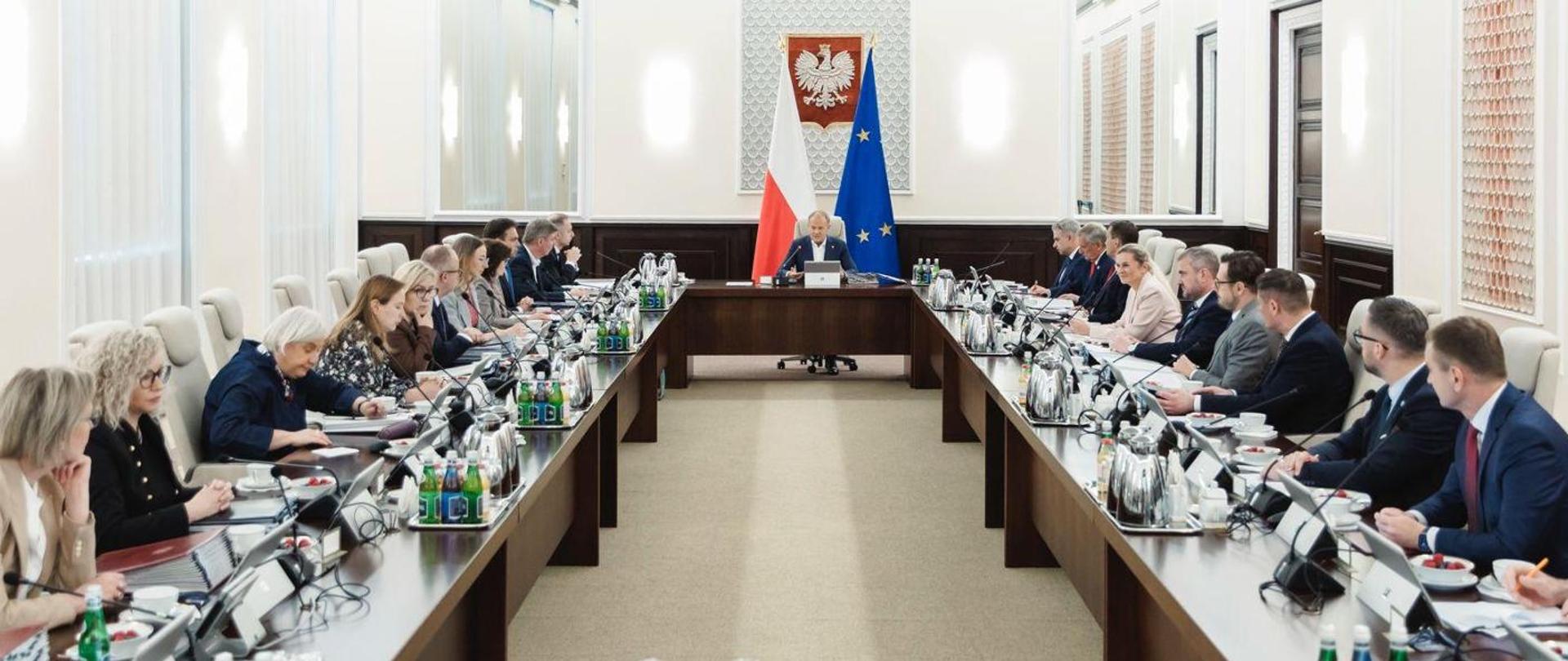 Sala posiedzeń rządu z długim stołem konferencyjnym; premier Donald Tusk siedzi pośrodku na czele stołu, otoczony przez ministrów i urzędników po obu stronach, w tle godło Polski oraz flagi Polski i Unii Europejskiej.