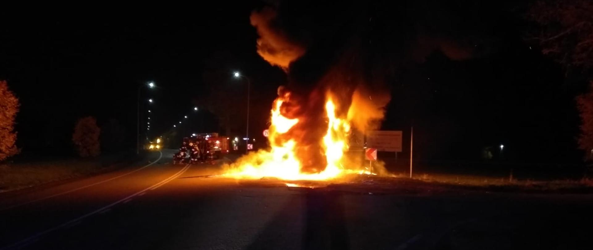 Zdjęcie palącego się samochodu dostawczego, w tle samochody strażackie i strażacy.