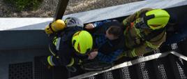 Ćwiczenia w DPS. Dwóch strażaków wyposażonych w sprzęt ochrony układu oddechowego ewakuuje po zewnętrznej klatce schodowej poszkodowanego mężczyznę, znajdującego się na wózku inwalidzkim. Strażacy mają żołte hełmy z czerwonymi paskami. 