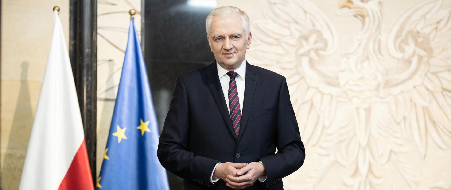 Jarosław Gowin stoi przodem do fotografa. W tle rysunek orła na ścianie i flagi polska i unijna.