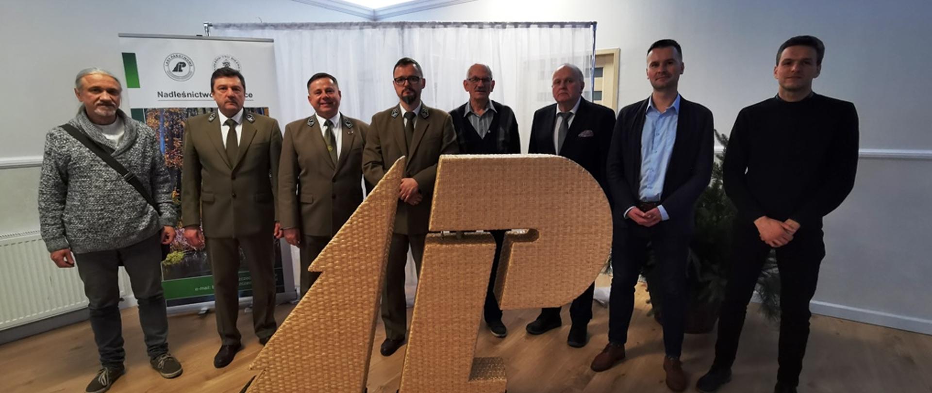 Grupa ośmiu mężczyzn pozuje do zdjęcia. Część z nich ubrana jest w zielone mundury leśników. Przed nimi stoją litery L i P, tworzące logo Lasów Państwowych.