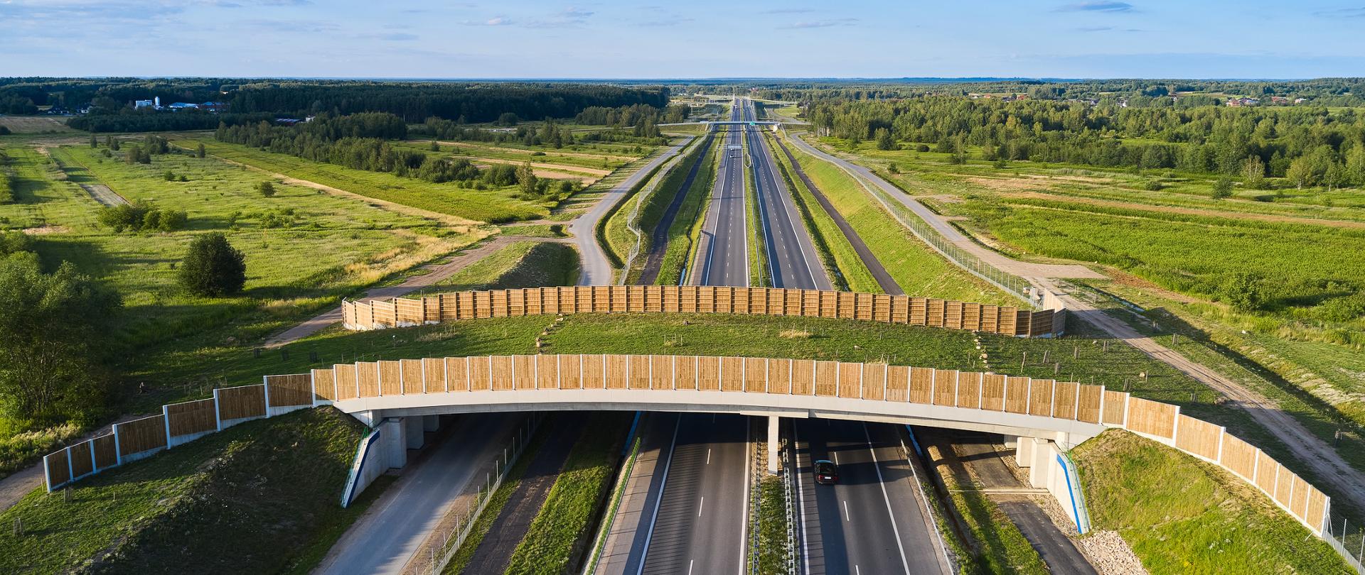 Widok z góry na drogę ekspresową. Na pierwszym planie widać przejście górne dla zwierząt. Dookoła lasy, świeci słońce. 