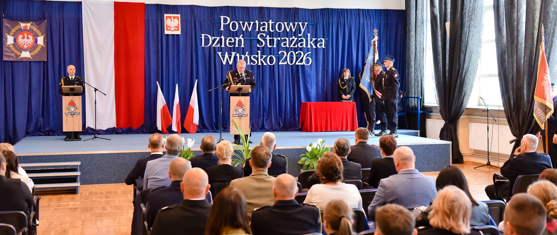 Zdjęcie przedstawia szeroki plan uroczystości z okazji Powiatowego Dnia Strażaka Wińsko 2026, odbywającej się w dużej sali. Widok obejmuje zarówno scenę z osobami prowadzącymi, jak i publiczność siedzącą tyłem do obiektywu. Główną część tła stanowi ciemnoniebieska kurtyna, na której widnieje duży, biały napis informujący o nazwie i dacie wydarzenia. Obok napisu wisi flaga Polski oraz godło państwowe. Na scenie znajdują się dwie mównice ozdobione logo straży pożarnej. Przy centralnej mównicy stoi mężczyzna w mundurze galowym, który przemawia do zgromadzonych. Przy drugiej mównicy, po lewej, stoi inny mundurowy. Po prawej stronie sceny stoi poczet sztandarowy ze sztandarem oraz strażacy w pełnym umundurowaniu galowym. W dolnej części zdjęcia widać rzędy krzeseł wypełnione przez publiczność. Wśród zgromadzonych są osoby w mundurach galowych, strojach koszarowych, piaskowych mundurach Ochotniczej Straży Pożarnej oraz osoby cywilne w strojach wizytowych.