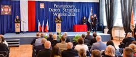 Zdjęcie przedstawia szeroki plan uroczystości z okazji Powiatowego Dnia Strażaka Wińsko 2026, odbywającej się w dużej sali. Widok obejmuje zarówno scenę z osobami prowadzącymi, jak i publiczność siedzącą tyłem do obiektywu. Główną część tła stanowi ciemnoniebieska kurtyna, na której widnieje duży, biały napis informujący o nazwie i dacie wydarzenia. Obok napisu wisi flaga Polski oraz godło państwowe. Na scenie znajdują się dwie mównice ozdobione logo straży pożarnej. Przy centralnej mównicy stoi mężczyzna w mundurze galowym, który przemawia do zgromadzonych. Przy drugiej mównicy, po lewej, stoi inny mundurowy. Po prawej stronie sceny stoi poczet sztandarowy ze sztandarem oraz strażacy w pełnym umundurowaniu galowym. W dolnej części zdjęcia widać rzędy krzeseł wypełnione przez publiczność. Wśród zgromadzonych są osoby w mundurach galowych, strojach koszarowych, piaskowych mundurach Ochotniczej Straży Pożarnej oraz osoby cywilne w strojach wizytowych.