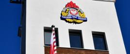 Flaga wisi na maszcie, za nią logo Państwowej Straży Pożarnej umieszczone na budynku.