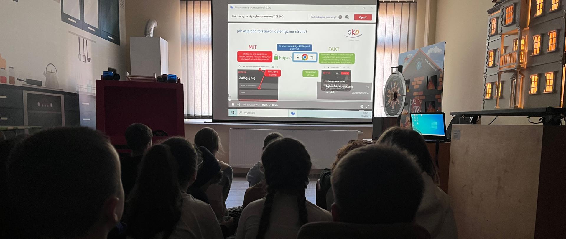 Na zdjęciu widać grupę dzieci siedzących na podłodze w ciemniejszym pomieszczeniu i oglądających prezentację wyświetlaną na ekranie. Tematem prezentacji wydaje się być cyberbezpieczeństwo, ponieważ widoczne są hasła dotyczące rozpoznawania fałszywych i autentycznych stron internetowych, a także oznaczenia „MIT” i „FAKT”.
Pokój ma wygląd przypominający klasę lub salę edukacyjną. Po prawej stronie widać makietę budynku z podświetlonymi oknami. Na ścianach znajdują się grafiki przedstawiające elementy kuchenne. W pomieszczeniu jest także komputer podłączony do monitora.
Scena wygląda na zajęcia edukacyjne, warsztaty dla dzieci na temat bezpieczeństwa w internecie.