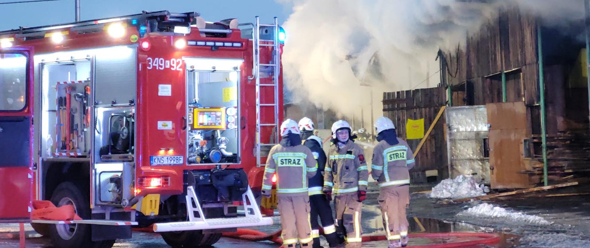 Samochód ratowniczo gaśniczy oraz strażacy obok płonącego budynku.