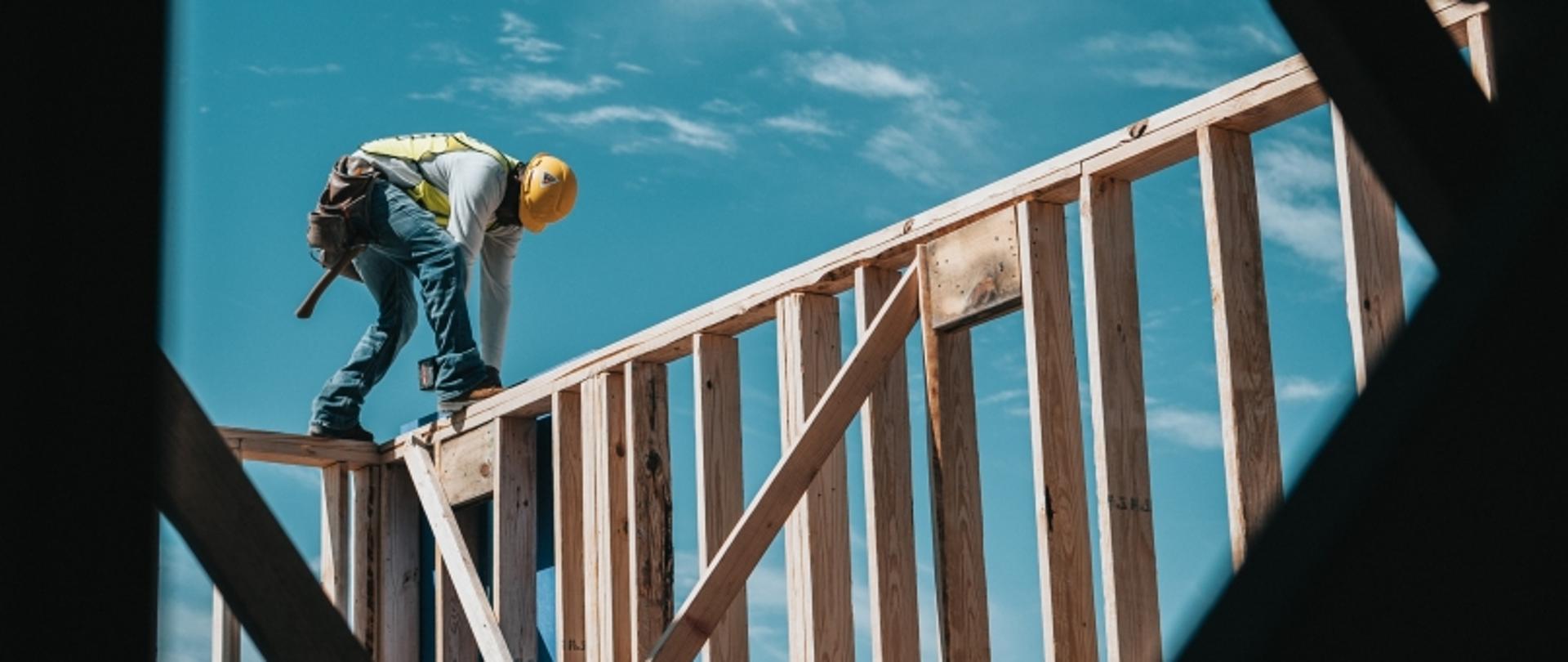 Budowlaniec w kasku i kamizelce pracujący na drewnianej konstrukcji budynku pod gołym niebem.