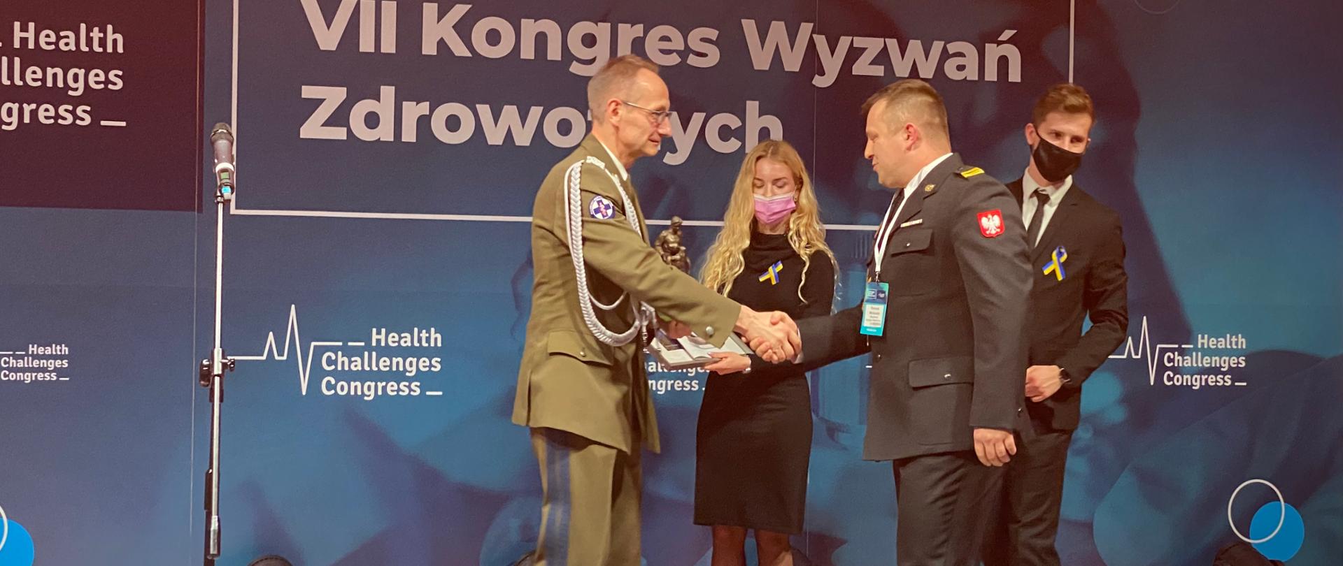 Podczas VII Kongresu Wyzwań Zdrowotnych generał wojska polskiego wręcza na scenie strażakowi nagrodę, asystuje mu dwie osoby kobieta i mężczyzną. W tle ścianka z napisem VII Kongres Wyzwań Zdrowotnych
