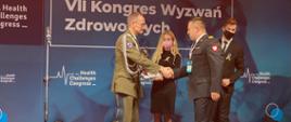 Podczas VII Kongresu Wyzwań Zdrowotnych generał wojska polskiego wręcza na scenie strażakowi nagrodę, asystuje mu dwie osoby kobieta i mężczyzną. W tle ścianka z napisem VII Kongres Wyzwań Zdrowotnych