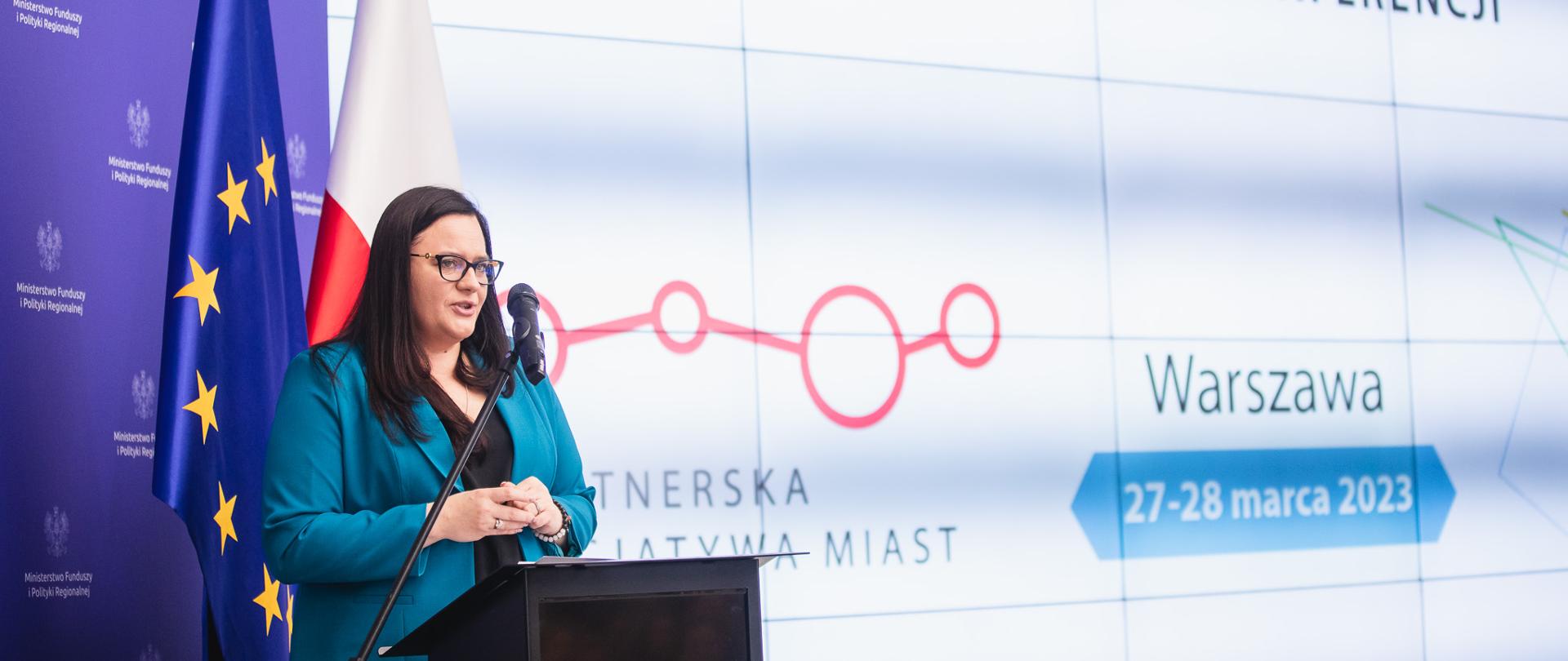 Wiceminister Małgorzata Jarosińska-Jedynak w sali konferencyjnej stoi w mównicy z mikrofonem. Za nią ekran z napisem: "Partnerska Inicjatywa Miast" oraz flagi Polski i UE.