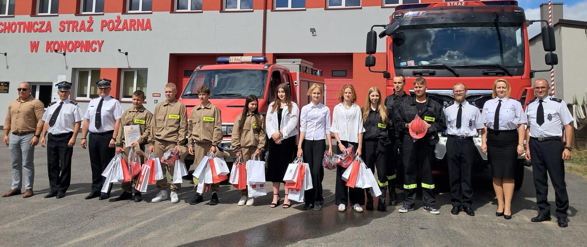 młodzież z MDP oraz druhowie i strażacy pozują do zdjęcia na tle samochodów pożarniczych i budynku jednostki