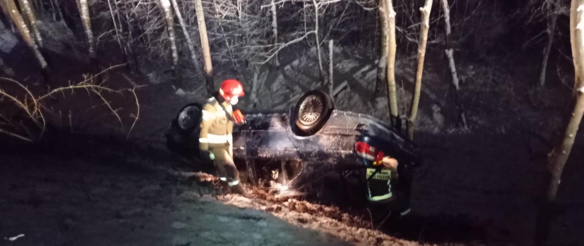 Samochód osobowy znajduje się na dachu w rowie. Obok niego stoi dwóch strażaków. 