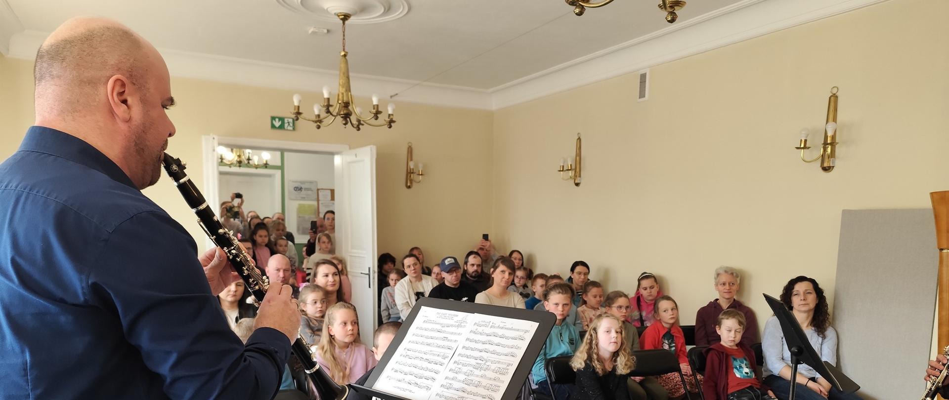 Nauczyciel grający na klarnecie, w tle widoczna publiczność.