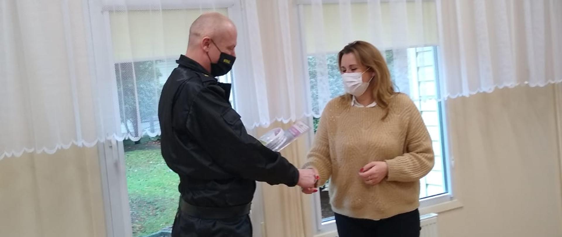 Zdjęcie przedstawia strażaka w ubraniu dowódczo-sztabowym oraz kobietę, którzy znajdują się w pomieszczeniu świetlicy. Strażak wręcza kobiecie czujkę tlenku węgla oraz dymu. Obie osoby mają na sobie maseczki chirurgiczne. 