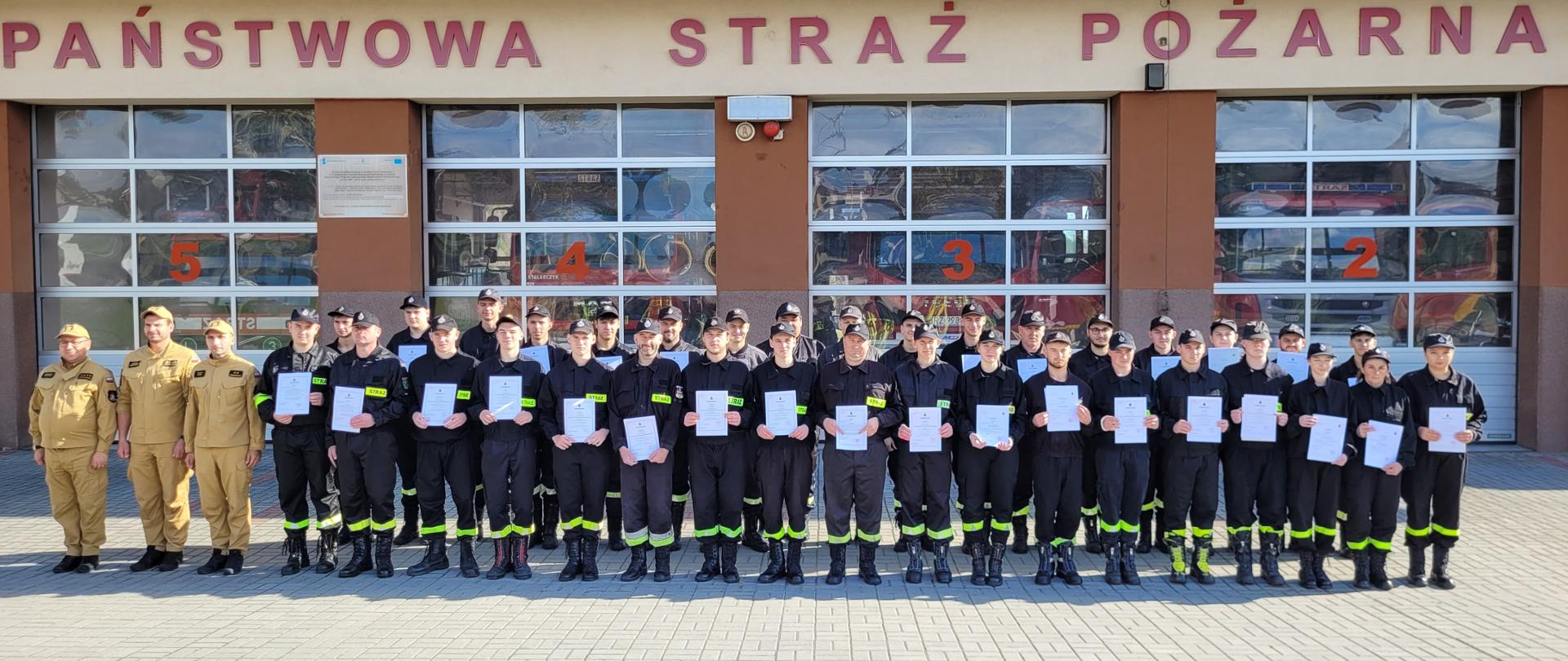 Na zdjęciu strażacy podczas zbiórki w dwuszeregu, w dłoniach trzymają zaświadczenia o ukończonym kursie podstawowym.