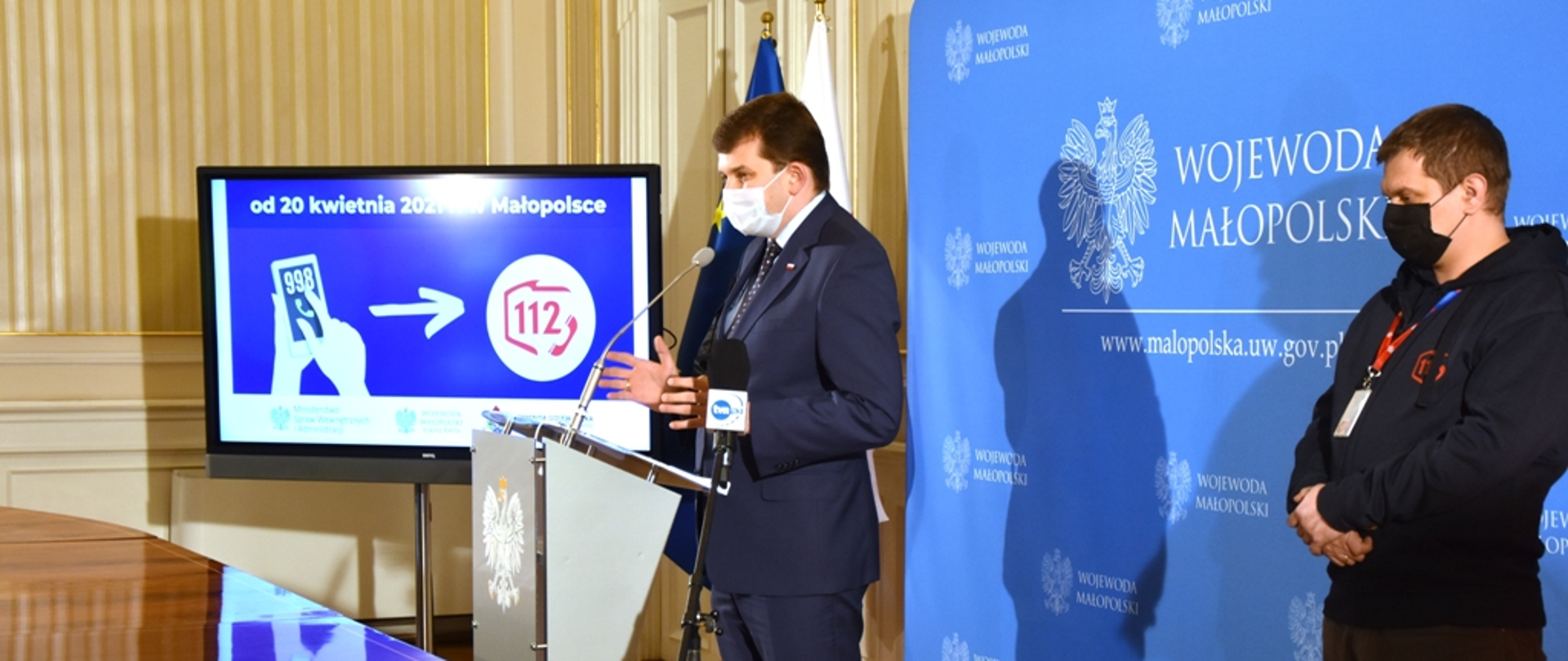 Zdjęcie obejmuje pomieszczenie w Małopolskim Urzędzie Wojewódzkim. Z prawej strony stoi kierownik CPR. W centralnym miejscu przy mównicy Wojewoda Małopolski informujący o przekierowaniu numeru alarmowego 998 do CPR. Z lewej strony telebim wyświetlający slajd.